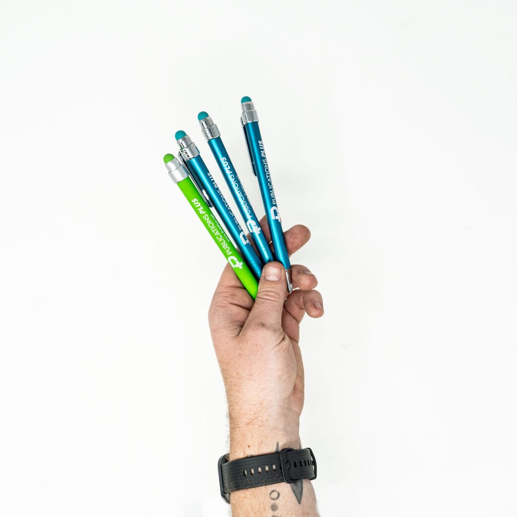 Hand holding green and blue pens that have a white logo printed on them.